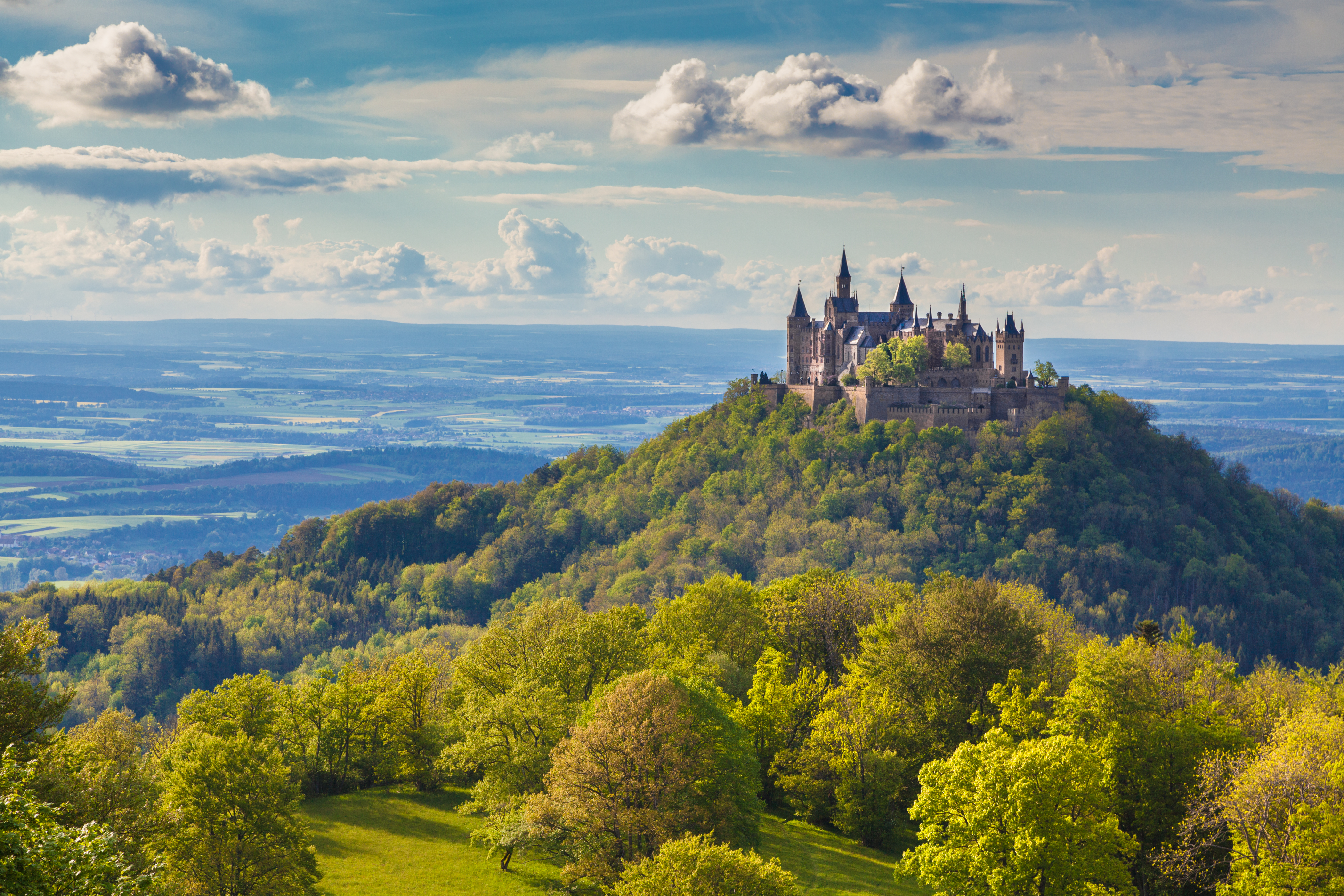Das Bild zeigt eine Fotoaufnahme der Burg Hohenzollern auf der schwäbischen Alb. Die Aufnahme ist vom Zeller Horn in der Nähe von Onstmettingen aufgenommen. Die Burg trohnt auf einem Hügel zwischen grünen Wäldern und Wiesen. Im Hintergrund ist der Zollernalbkreis und ein strahlend blauer Himmel zu sehen. 